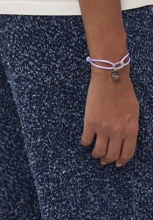 Purple bracelet with double cord design and silver accents, featuring a round charm marked "studios." Hand with dark skin against textured blue pants.