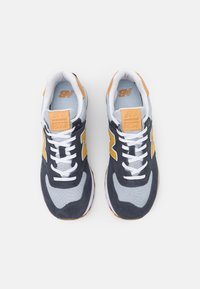  Paire de baskets New Balance 574 en gris, bleu marine et jaune, avec des lacets blancs et des patchs logo beige sur les languettes.