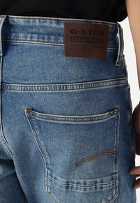 El bolsillo trasero de unos jeans de mezclilla azul con una etiqueta de cuero marrón de la marca G-Star RAW Originals encima.