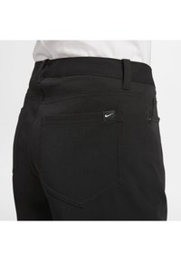 Close-up of black pants with visible back pocket and small white Nike swoosh logo on the pocket tag, worn with a white shirt.