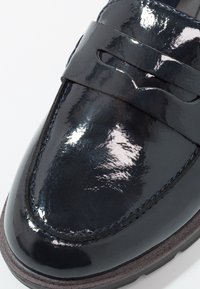 Chaussure en cuir verni bleu marine au design élégant, avec une large lanière sur le dessus et des coutures visibles le long du bord de la semelle.