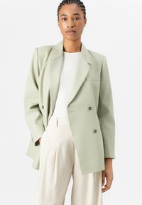 Dobbeltbrettet blazer i lysegrønt stoff med store revers, frontlomme og svarte knapper; båret over en hvit ribbestrikket topp.