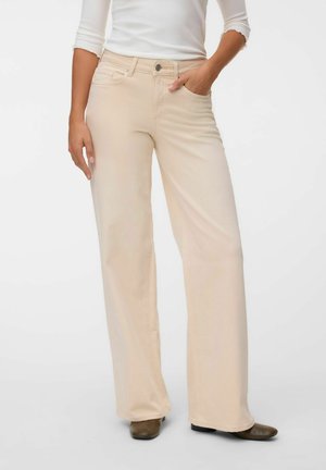 Femme portant un pantalon beige taille haute à jambes larges, un haut blanc à manches longues, une main dans la poche et des chaussures marron à talons bas visibles.