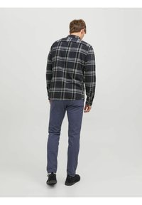 Uomo di spalle che indossa una camicia a quadri neri e grigi, pantaloni blu e scarpe nere, su uno sfondo bianco uniforme.