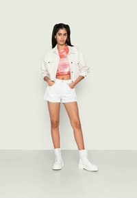 Jaqueta de ganga branca sobre um top cropped rosa tie-dye, combinada com calções brancos e botins brancos texturizados. Modelo de pé contra um fundo neutro.