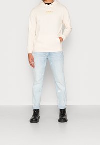 Beige hoodie med framficka och dragsko på huvan, ihop med ljusblå slitna jeans och svarta snörskor.