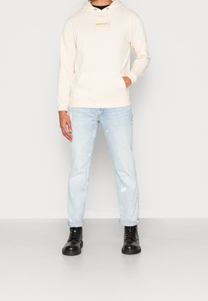 Felpa beige con tasca frontale e cappuccio con stringhe, abbinata a jeans chiari strappati e stivali neri con lacci.
