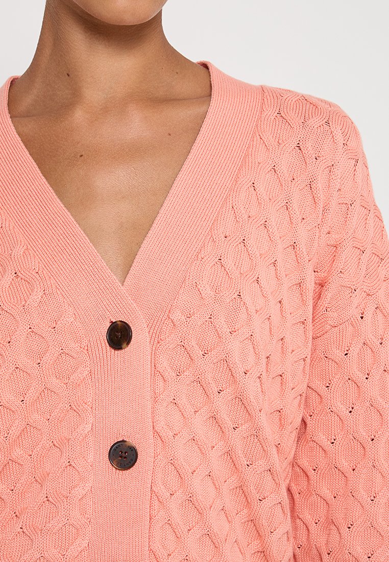 Sweter z dzianiny w miękkim różowym kolorze z dekoltem w kształcie litery V, z ozdobnym wzorem w romby i dwoma brązowymi guzikami z przodu.