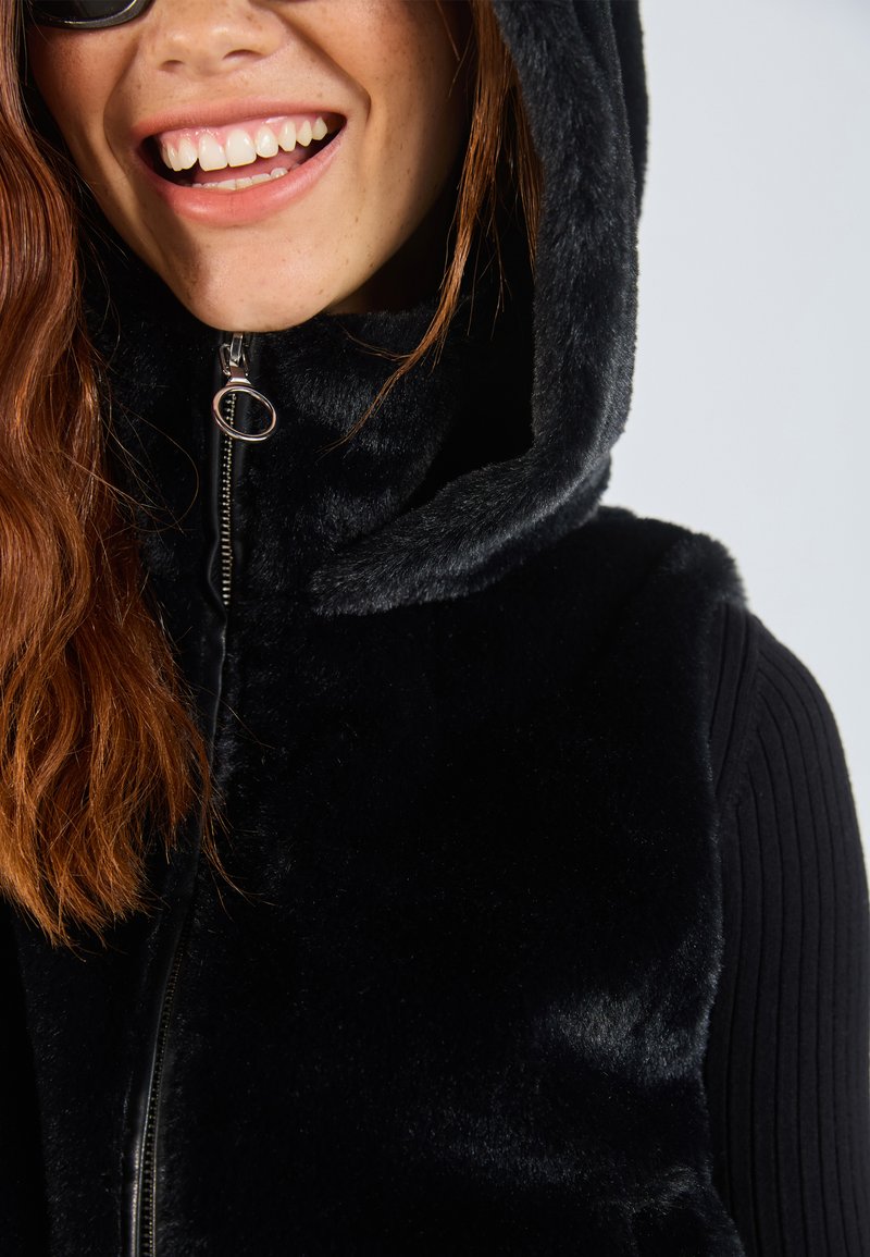 Gilet in pelliccia sintetica nera con cappuccio e chiusura con zip; presenta una texture morbida e cuciture a outline. Indossato sopra una maglia nera a costine.