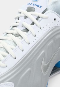 Nike Shox tenisice u bijeloj boji sivi akcenti, s glatkom teksturom, perforiranim detaljima i zaobljenim vezicama. Plavi brend na jeziku.