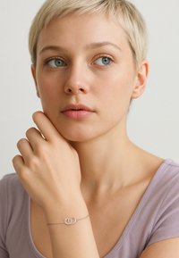 Silverarmband med sammanflätade cirkulära länkar, bärs på en hand som vilar mot hakan, mot en neutral bakgrund.