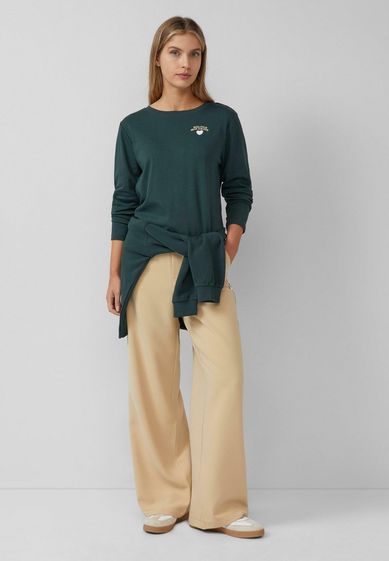 Donkergroen langemouwen shirt met een klein wit logo, gecombineerd met lichtbeige wijde broek en crèmekleurige schoenen met een gestructureerde zool.