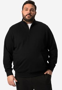 Sudadera negra con cremallera en un tejido suave y texturizado, con un bolsillo frontal y puños acanalados, llevada sobre una camisa blanca. Diseño simple y casual.