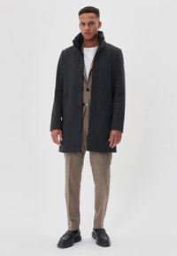 Abrigo gris oscuro con un cuello alto, cierre de botones y bolsillos laterales, llevado sobre un traje claro y pantalones beige, combinado con zapatos negros.