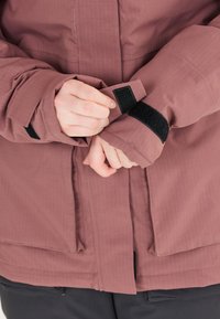 Locker sitzende, mauvefarbene Jacke mit strukturiertem Finish, ausgestattet mit schwarzen Klettverschlussmanschetten und einem Reißverschluss vorn. Enthält einen leichten Glanz und Seitentaschen.