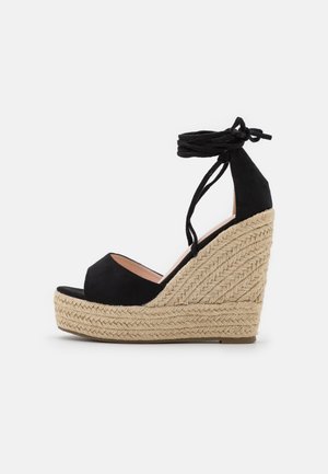 Czarna sandałka na koturnie zamszowa z grubą, plecioną platformą z juty i wiązaniami wokół kostki, pokazana z boku na białym tle.