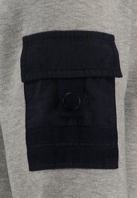 Tessuto grigio con una tasca a toppa blu navy scura, caratterizzata da una chiusura a bottone a pressione e due bande orizzontali, che aggiungono texture e contrasto.