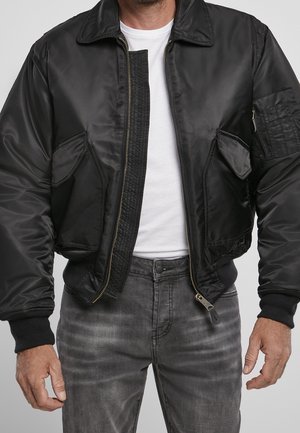 Homme portant une veste bomber noire sur un t-shirt blanc avec un jean gris, debout, les mains détendues le long du corps.