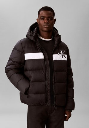 Junger Mann trägt eine schwarze Steppjacke mit einem weißen horizontalen Streifen und "cK"-Logo, steht vor einem einfarbigen grauen Hintergrund.