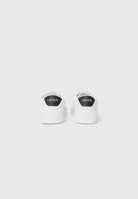 White sneakers with a textured upper, featuring a black Lacoste logo at the heel, and a white rubber sole. Minimalist design.