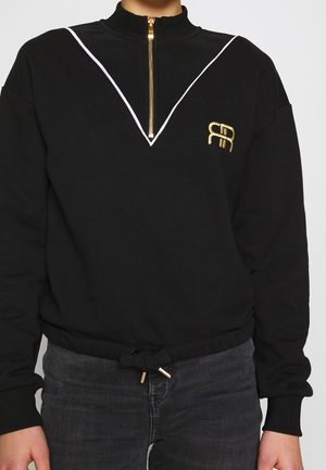 Pull-over noir à manches longues avec fermeture éclair dorée, bordure blanche en forme de V, logo doré brodé sur la poitrine, et taille à cordon, porté avec un jean foncé.