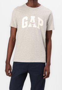 Mand iført en lysegrå T-shirt med korte ærmer og teksten "GAP Original" samt mørkeblå cargo-bukser, står imod en hvid baggrund.