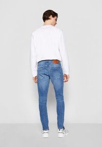 Jeans skinny de color azul claro con un ligero desvanecimiento, cinco bolsillos y un parche de cuero marrón en la cinturilla, combinados con una camisa blanca de manga larga.