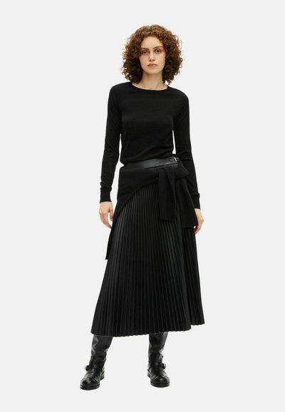 Maglione nero a maniche lunghe abbinato a una gonna plissettata nera accentuata da una cintura nera. Il materiale ha una trama liscia. Stivali neri al ginocchio.