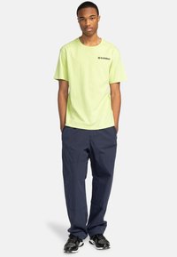 Neon grøn bomulds t-shirt med sort logo, parret med marineblå cargo-bukser og sorte træningssko. Casual, afslappet pasform, korte ærmer.