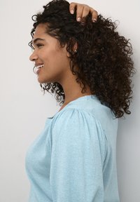 Maglione azzurro chiaro con corpo strutturato e maniche a sbuffo, indossato da una persona che sorride, capelli acconciati in riccioli morbidi, su uno sfondo bianco semplice.