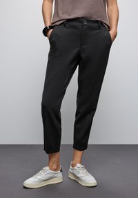 Pantalons noirs ajustés avec une texture lisse, dotés d'une fermeture à boutons à l'avant et de poches latérales, associés à des baskets de couleur claire.