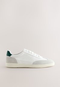 Baskets blanches basses avec des accents en daim gris, languette de talon verte et lacets blancs sur un fond neutre, vue de côté.