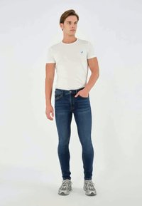 Smal måte passform mørkeblå denimbukser kombinert med en enkel hvit t-skjorte. Modellen har på seg grå joggesko. Enkel design med minimal detaljer.