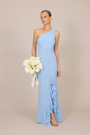 Žena v svetlomodrých asymetrických šatách držiaca biele kala kvety, nosiaca zlaté sandále a výrazné náušnice na jednofarebnom béžovom pozadí.