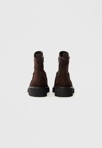 Une paire de bottes en daim marron foncé avec des semelles noires épaisses, vues de l'arrière sur un fond gris clair uni.