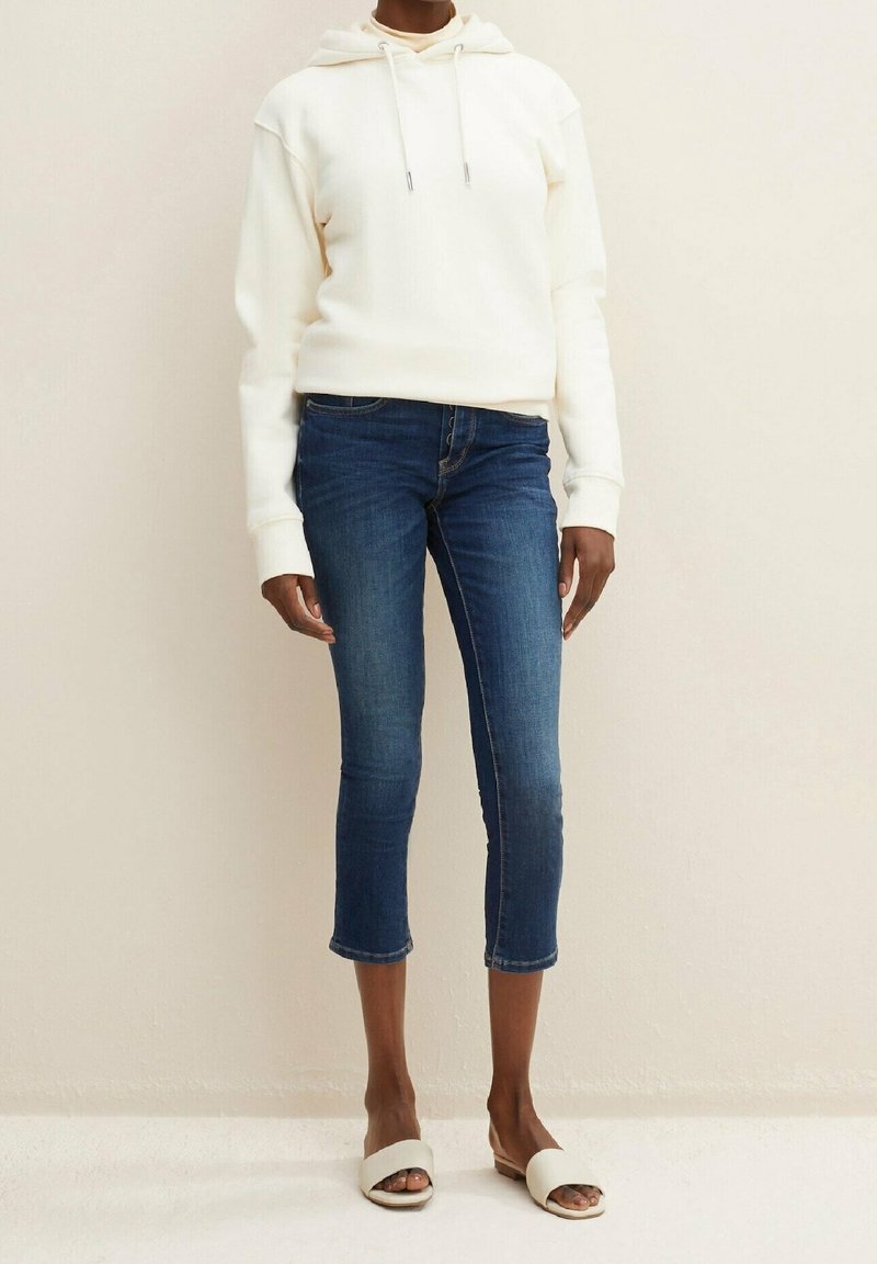 Vit hoodie med snörning, i kombination med mörkblå korta jeans och öppna vita sandaler. Slät tyg och avslappnad passform.