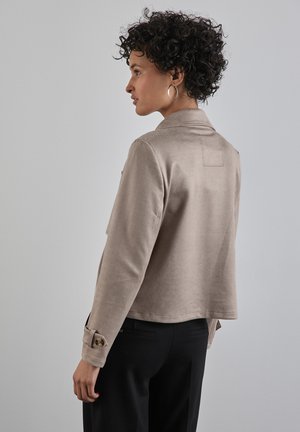 Veste en velours côtelé beige avec un col à revers, des poches avant et des poignets boutonnés, associée à un pantalon noir. Tissu texturé et coupe décontractée.