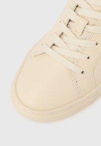 Közeli kép egy bézs tornacipőről, krém színű fűzőkkel, látható varrással és texturált talppal, fehér háttéren.