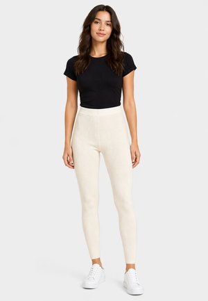 Frau steht und trägt ein eng anliegendes schwarzes Kurzarm-Top, hoch taillierte cremefarbene gerippte Leggings und weiße Sneakers auf einfarbigem Hintergrund.