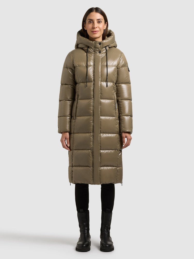 Abrigo acolchado en verde oliva, con capucha, bolsillos con cremallera y diseño acolchado. Combinado con pantalones ajustados negros y botas negras gruesas.