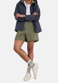 Námořnická voděodolná bunda s kapucí přes bílé tričko, v kombinaci s olivově zelenými šortkami, pruhovanými ponožkami a béžovými teniskami.