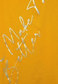 Žlutá tkanina s metalickým stříbrným kurzívním textem "Make Better" v diagonálním uspořádání.