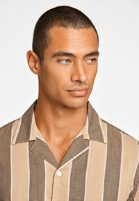 Camisa de manga corta con cuello en rayas beige y marrón; cuenta con un cierre de botón y una tela texturada con un ajuste relajado.