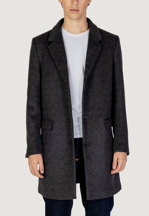 Cappotto di lana grigio con colletto a rever, due tasche frontali e chiusura a bottoni, indossato sopra una maglietta bianca e jeans scuri.