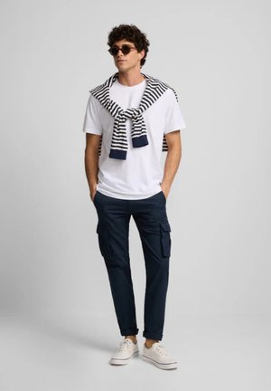 Conjunto informal que incluye una camiseta blanca, pantalones cargo azul marino y un suéter a rayas atado sobre los hombros. Los accesorios incluyen gafas de sol y zapatillas blancas.