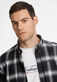 Camicia di flanella a quadri in bianco e nero con colletto button-down, abbinata a una maglietta bianca con un logo blu e rosso.