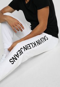 Białe spodnie dresowe z czarnym napisem "CALVIN KLEIN JEANS" wzdłuż lewej nogawki. Wykonane z miękkiej tkaniny, o luźnym kroju i elastycznym pasie.