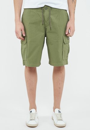 Uomo che indossa pantaloncini cargo verde oliva con vita con bottoni e coulisse, tasche laterali, sneakers bianche e una camicia bianca infilata dentro.