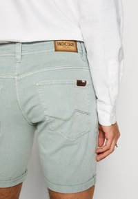 Homme portant un short en denim vert clair avec des poches arrière et un patch de marque en cuir, associé à une chemise blanche à manches longues boutonnée.