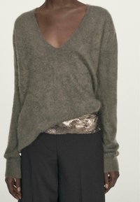 Olivgrüner, gestrickter Pullover mit tiefem V-Ausschnitt und langen Ärmeln, verziert mit schimmernden Pailletten am Saum, kombiniert mit dunklen Hosen.